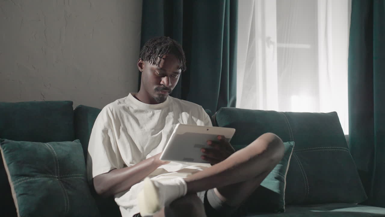 Champ sitting comfortably on teal sofa, focused on tablet in peaceful living room with natural light. Quiet, serene moment captured in stylish, modern home setting with soft shadows