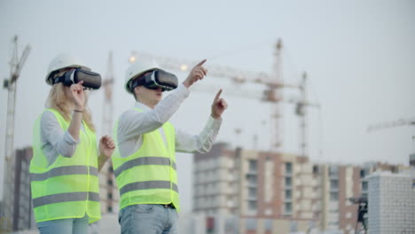 Two-people-Man-and-woman-wearing-virtual-reality-glasses-on-the-background-of-buildings-under-construction-with-cranes.-Manager-and-assistant-on-design-with-your-hands-mimicking-the-interface-of-the-application