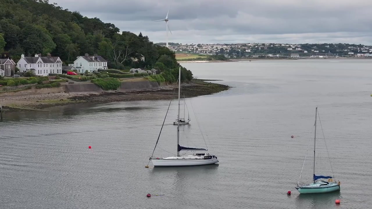 Et roligt luftperspektiv af sejlbåde forankret i havnen ved Crosshaven, County Cork, med kysthuse og en vindmølle under en overskyet himmel