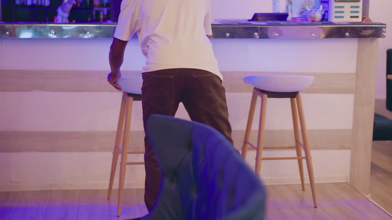 Man adjusting white stool at modern bar in stylish purple-lit living room, preparing space for social gathering or event, with focus on cozy, trendy furniture in elegant interior setting
