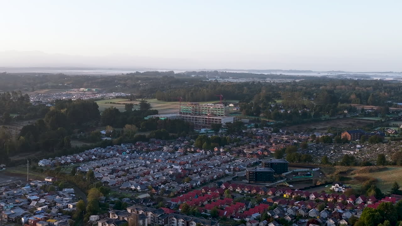 Luftaufnahmen einer Drohne bei Sonnenaufgang, die die Baustelle des zukünftigen Krankenhauses in Puerto Varas, Chile, mit Kränen und Morgenlicht zeigen.