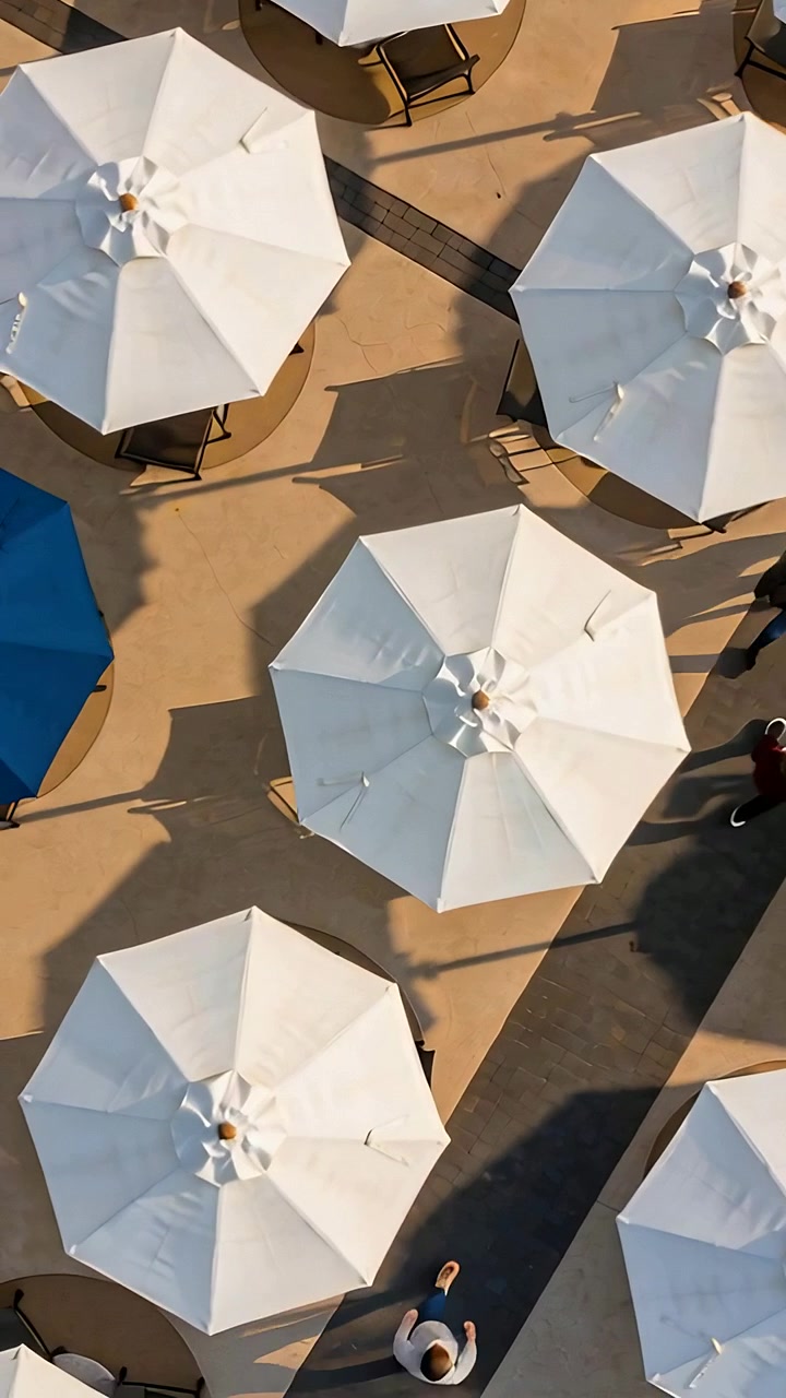 Video verticale: Drone in panoramica che mostra una griglia di ombrelloni in una piazza, pedoni che attraversano oltre un tendone blu.