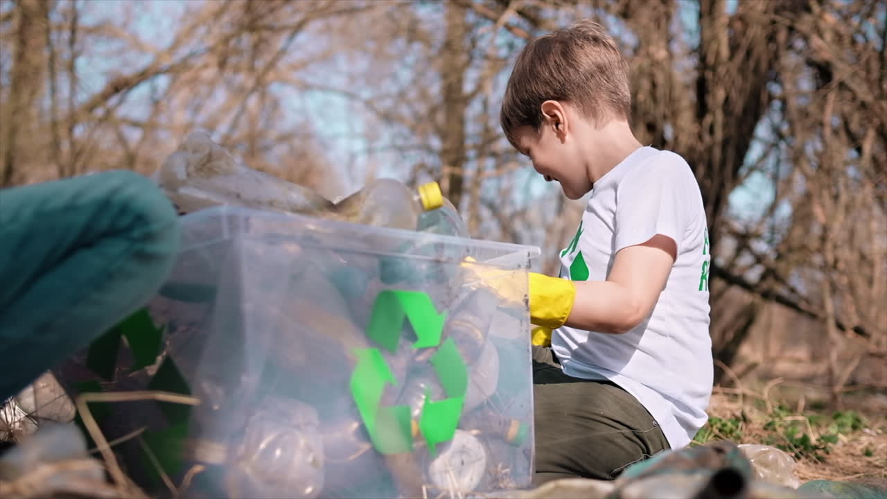 Boy and girl collecting plastic garbage in a container in a polluted clearing, recycling signs on the T-shirts. Slow motion