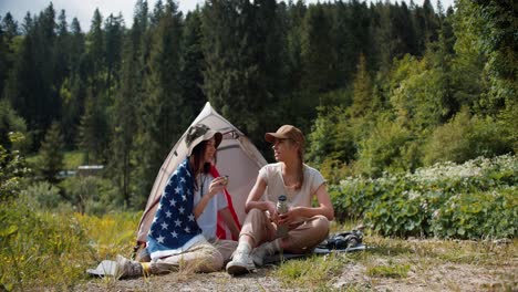 A-blonde-girl-and-a-brunette-girl-in-a-hat-who-swam-in-the-American-flag-sit-near-the-tent-against-the-backdrop-of-a-green-forest-and-communicate