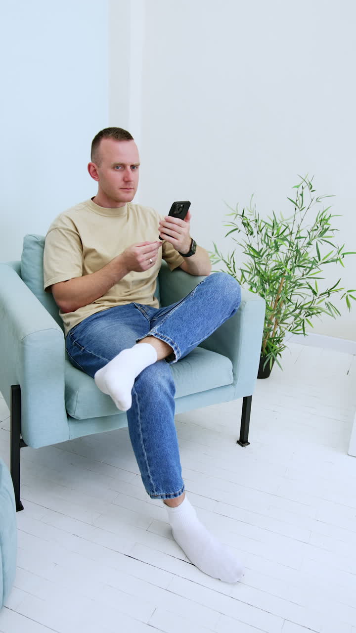 Man wearing beige t-shirt and jeans sits comfortably in the armchair indoors. Man looks at his phone expressing surprise. Vertical video.