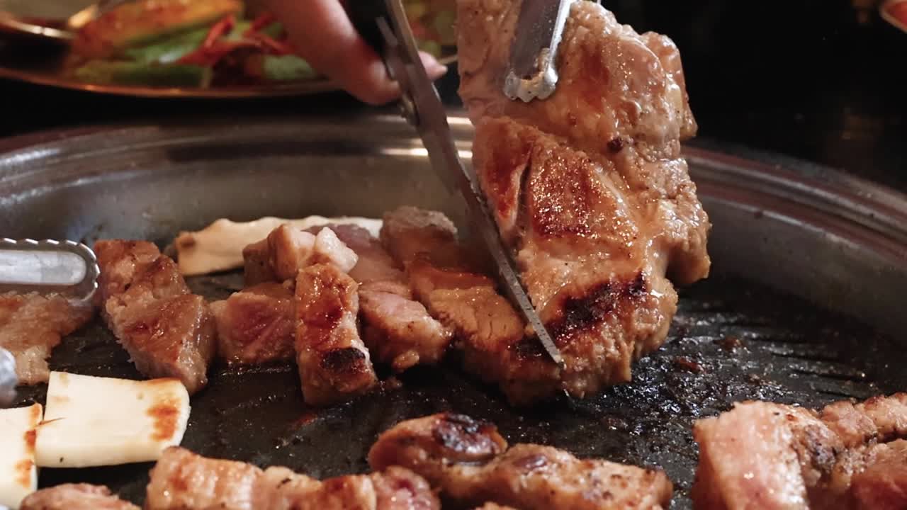 Vue en gros plan de mains utilisant des ciseaux et des pinces métalliques pour découper du porc grillé grésillant.