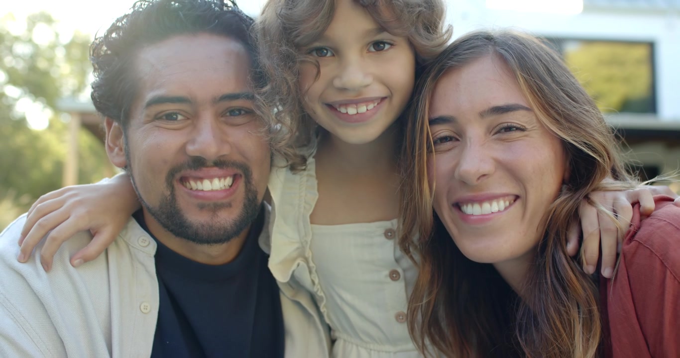 Familia diversa agachándose y acercándose, niño/a entre los adultos en el porche, sonriendo para el teléfono.
