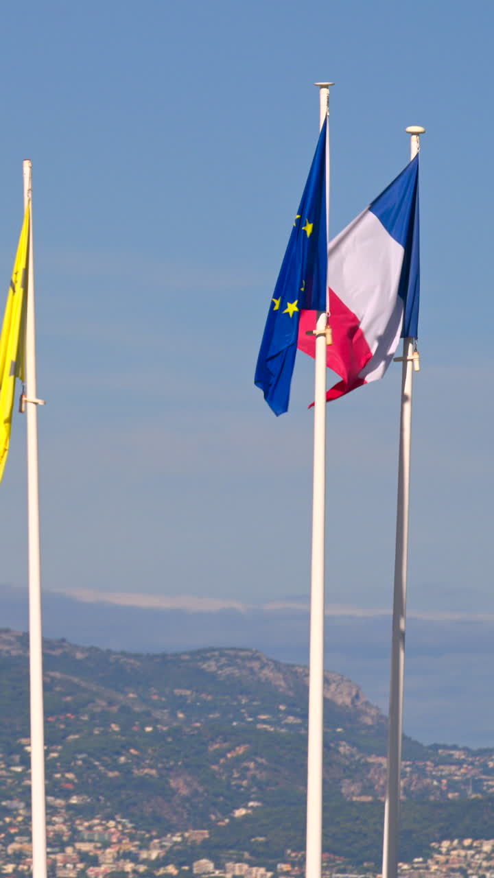 Ranskan lippu, Euroopan unionin lippu ja Villeneuve-Loubet’n lippu liehuvat tuulessa, ja taustalla näkyvät vuoret Ranskassa. Pystykuva
