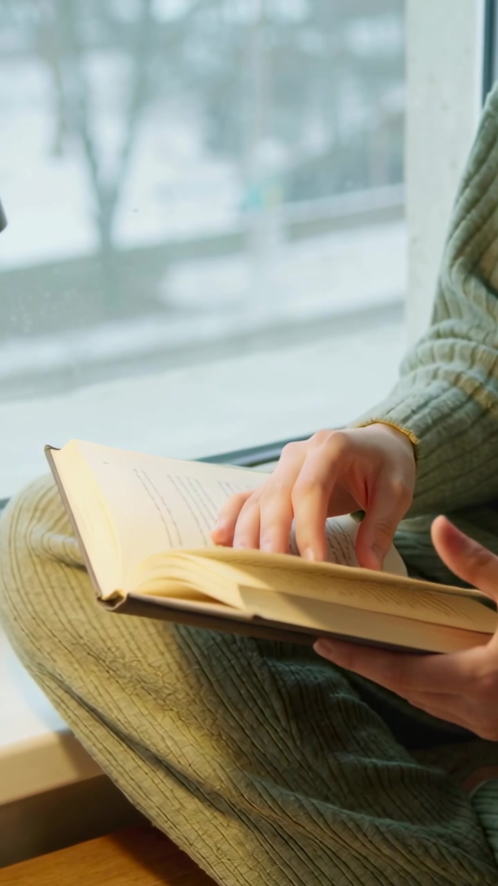 Video verticale: Donna con maglione verde che si gira, lisciando le pagine, seduta al finestrino leggendo un libro aperto.