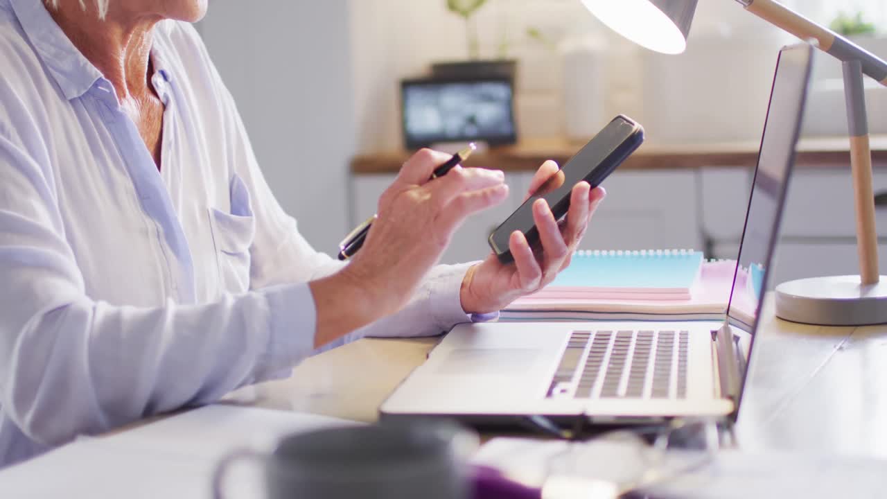 노인 백인 여성이 부 ⁇ 에서 테이블에 앉아 노트북과 스마트폰을 사용하고 있습니다.