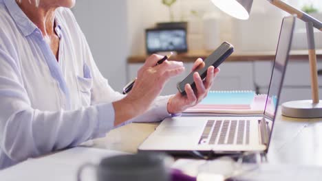mujer caucásica mayor sentada en la mesa en la cocina, usando computadora portátil y teléfono inteligente