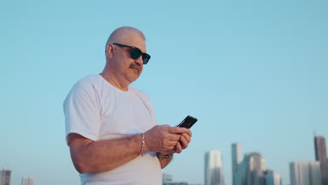 Low-angle-view-of-attractive-business-man-over-blurred-street-background-holding-smart-phone-in-hands-using-4G-internet-chatting-with-friends-searching-contact.-Electronic-wireless-device-concept.