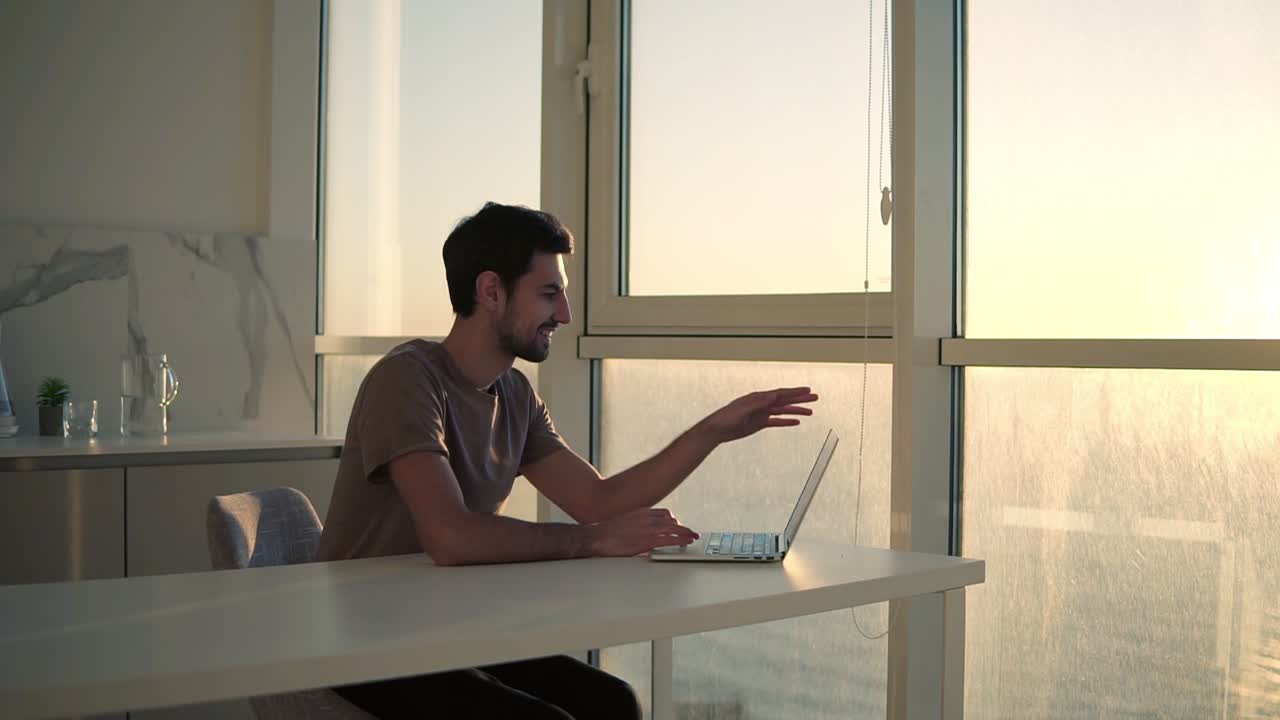 European dark haired man sitting close to panoramic window on studio kitchen - looking on laptop deskop and cheerfully smiling, chatting with friends. Slow motion. Side view