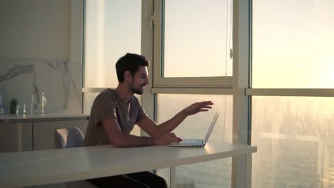 european dark haired man sitting close to panoramic window on studio kitchen - looking on laptop deskop and cheerfully smiling, chatting with friends. slow motion. side view