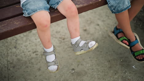 las piernas de dos niños pequeños mientras están sentados en un banco, un niño tiene la pierna cruzada, y el otro está balanceando suavemente su pierna derecha. ambos llevan sandalias