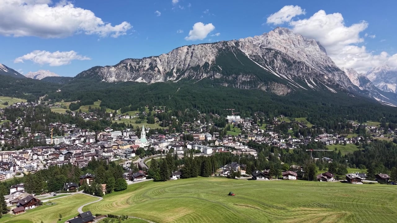 Góc nhìn từ flycam trên không qua Cortina d'Ampezzo, với thị trấn và những đồng cỏ núi cao xung quanh được bao bọc bởi những vách đá vôi hùng vĩ của Núi Pomagagnon dưới bầu trời mùa hè xanh trong