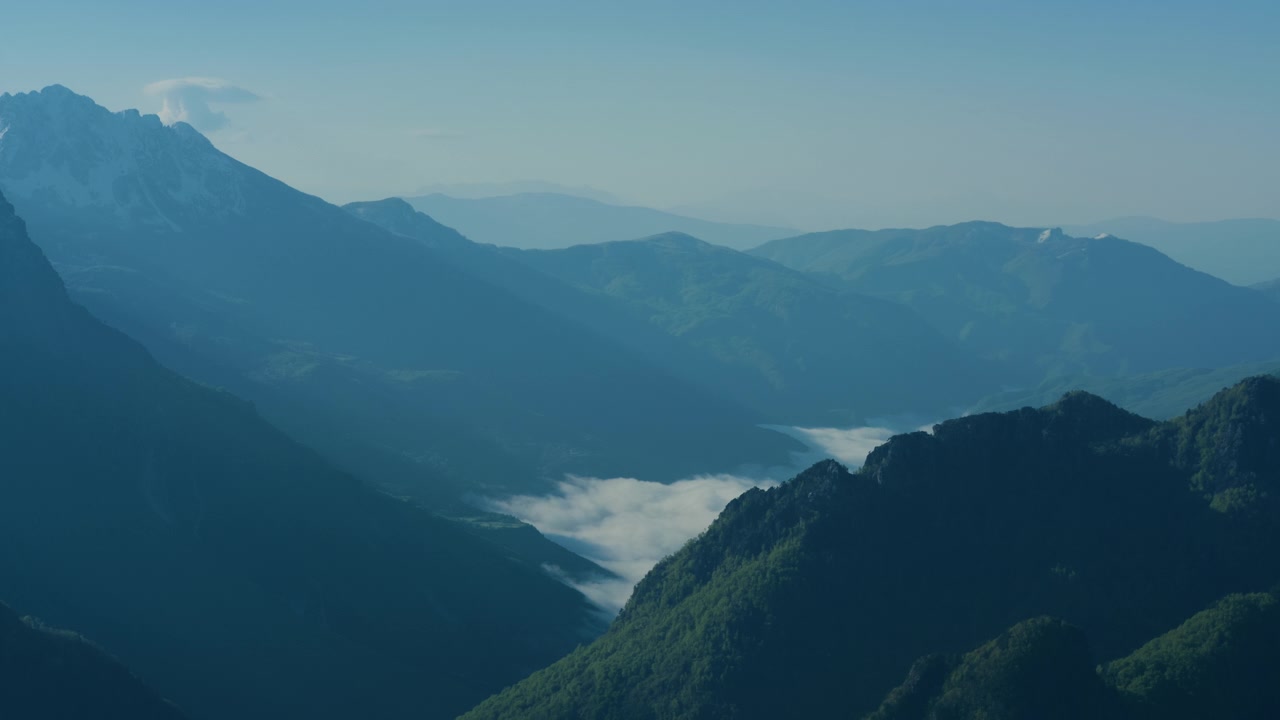 低い雲と青い霧に包まれた山間の谷、静止した俯瞰の風景。澄み切った空の下、美しい緑の牧草地を見下ろす雪を頂いた高山の峰々。