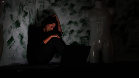 depressive young woman with tape over mouth reading bad news or hate messages on laptop in dark corner of room