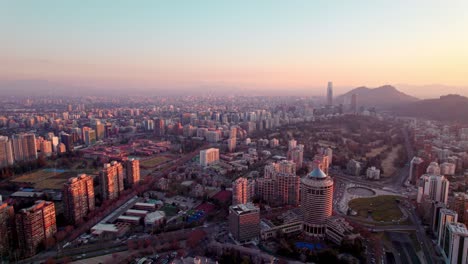 Aerial-orbit-of-modern-Santiago-buildings-and-skyscrapers,-mountains-in-back-at-golden-hour,-Chile