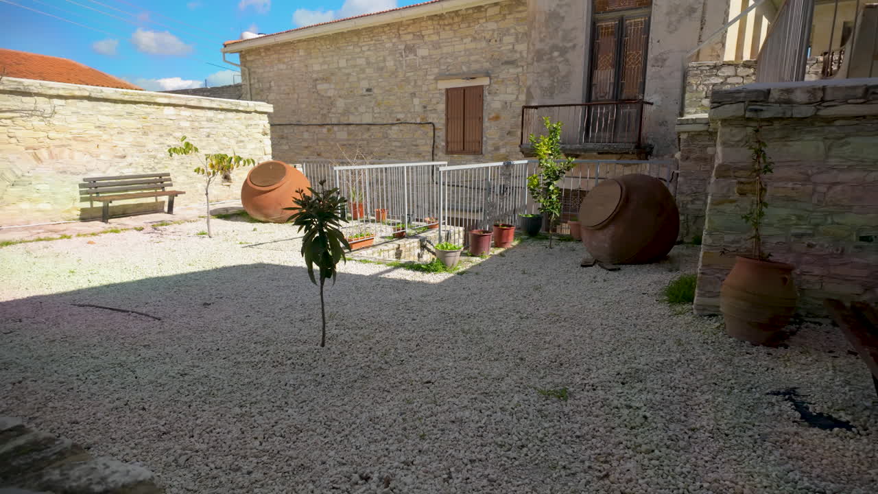 A peaceful stone courtyard featuring gravel, potted plants, and large clay jars, flanked by traditional stone buildings in Lefkara
