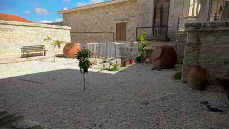 un pacífico patio de piedra con grava, plantas en macetas y grandes jarras de arcilla, flanqueado por edificios tradicionales de piedra en lefkara