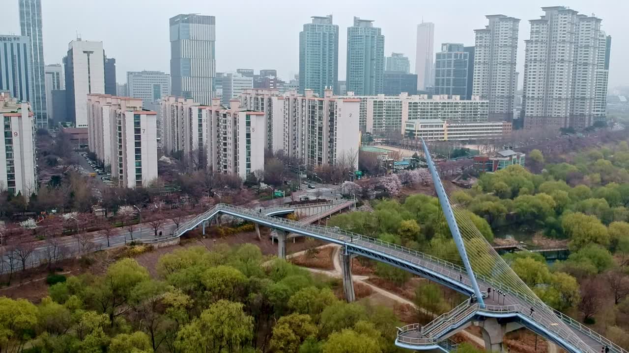 A forward motion shot of an artistic bridge over a green space, with a cityscape of skyscrapers visible in the background