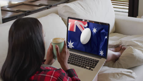 biracial woman watching laptop with rugby ball on flag of australia on screen