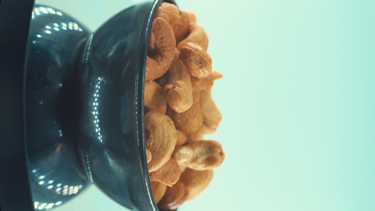 Una ripresa verticale ravvicinata di un anacardo salato tostato in una ciotola nera lucida su un supporto rotante, in slow motion, 4k.