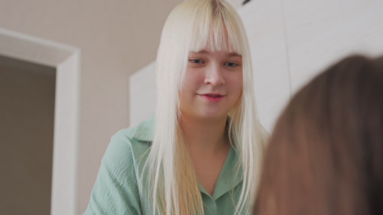 blonde girl smiles warmly while interacting with brunette friend during playful indoor bonding moment, expressing joy and emotional connection in relaxed home environment with natural lighting