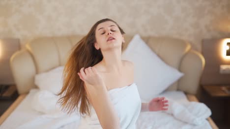 bella chica sentada en la cama se quita una toalla de la cabeza y se suelta el pelo