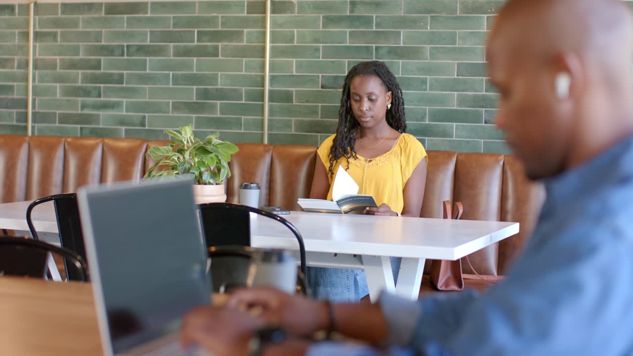 African American patrons, man working on laptop while woman in yellow top reading book at cafe