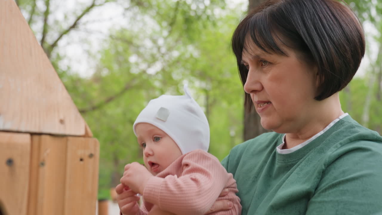 Grandmother Plays Outdoors, Ancient Dame Demonstrates Wooden Toy Enthusiastically Outdoors, Elderly Grandmother Engaging And Supporting Infant With Handcrafted Wooden Toy In Vibrant Park Setting