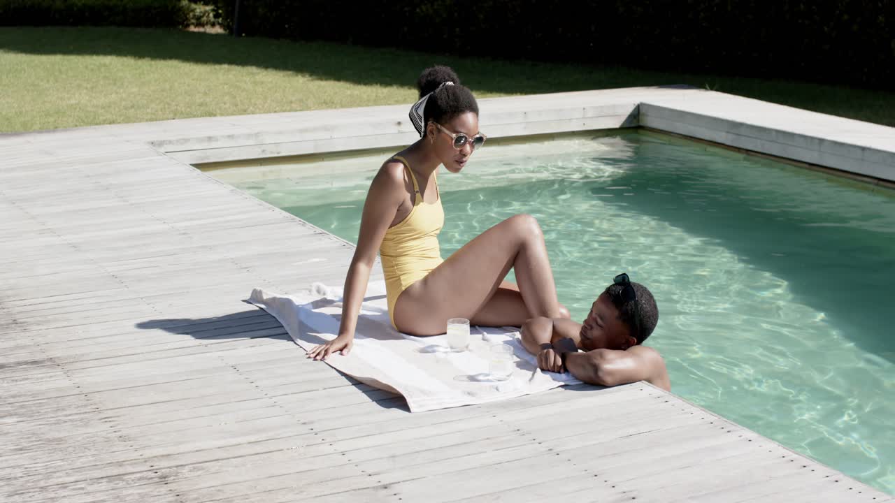 Afroamerikanisches Paar unterhält sich am Pool, Frau im gelben Badeanzug greift nach einem Glas auf einem Handtuch; mit Freiraum für Text.