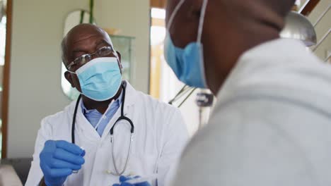 african american senior male doctor giving covid test to male patient in home, wearing face masks