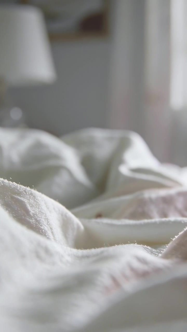Video vertikal: Kamera bergeser memperlihatkan lipatan selimut kusut di tempat tidur untuk menunjukkan tekstur dalam cahaya lembut.