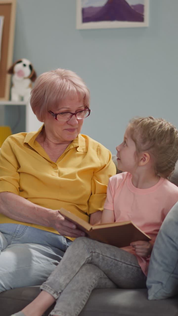 Naughty blonde little child reads book with grandmother on sofa in living room slow motion. Boring lessons at home. Elderly woman and girl do homework