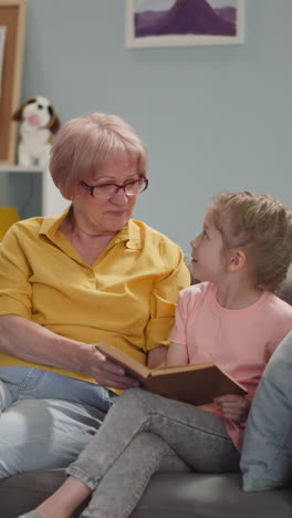 naughty blonde little child reads book with grandmother on sofa in living room slow motion. boring lessons at home. elderly woman and girl do homework