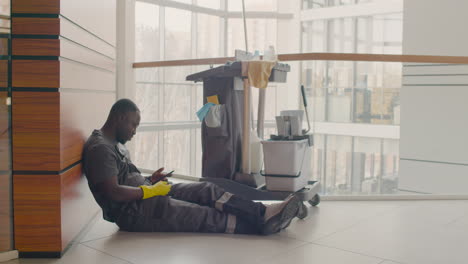 cleaning man sitting on the floor while looking at the mobile near a cleaning cart