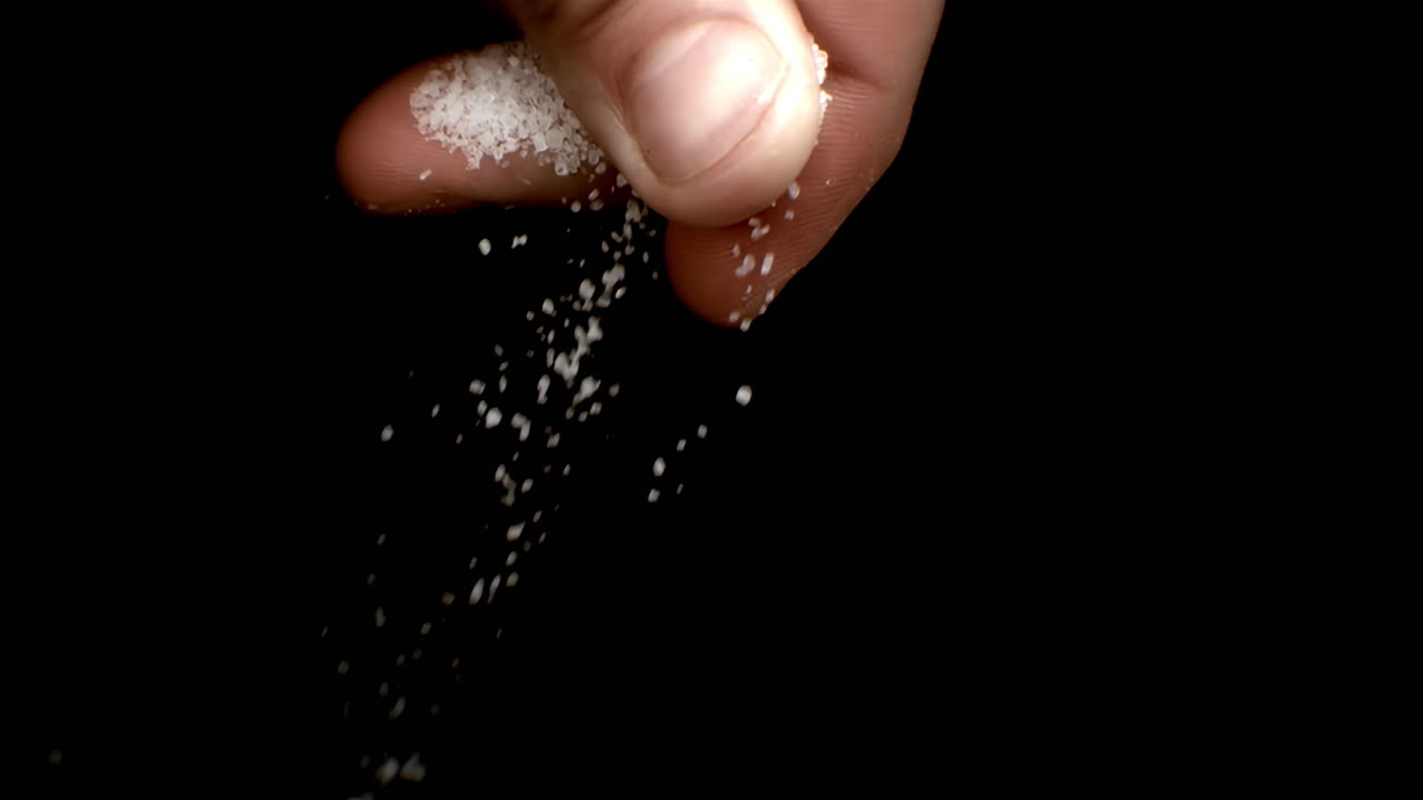 Fingers sprinkling fine salt grains over dark surface in slow, close up motion
