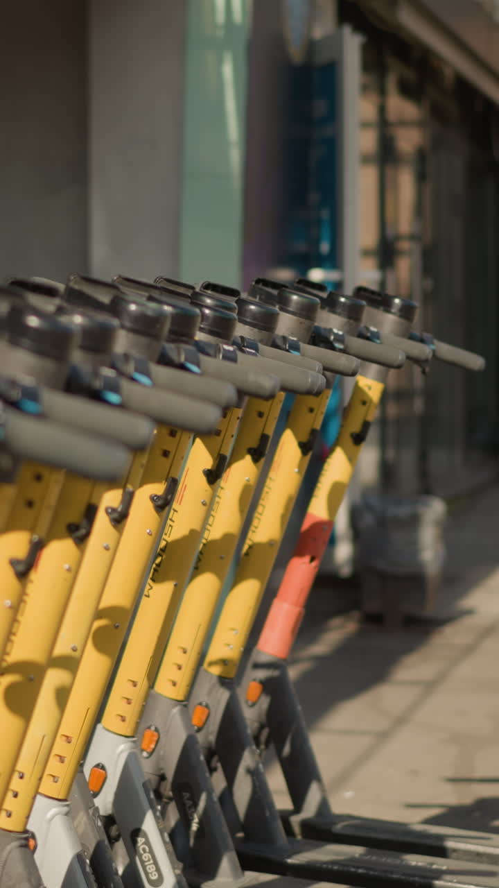 노란색 전동 스쿠터가 인도에 줄지어 있고, 아침 햇살이 부드러운 그림자를 드리우고 있어요. 깔끔하게 정돈된 핸들바와 브레이크 레버, 도시 경계의 눈에 띄는 노란색 프레임, 통근자들을 기다리는 대여 차량들이 보이네요.