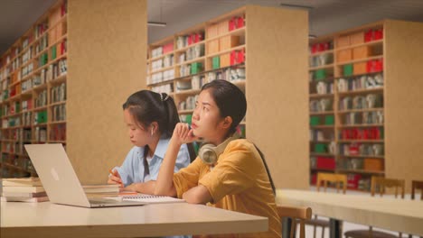 asian woman student with headphones thinking while sitting with her classmate writing into the notebook on a table with a laptop in the library