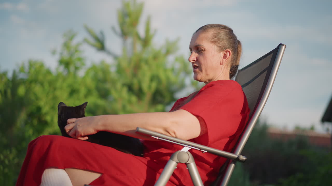 Caucasian woman in red dress resting on reclining garden chair holding black cat, suburban backyard greenery, soft daylight, calm expression, gentle petting, slow living atmosphere, therapeutic calm