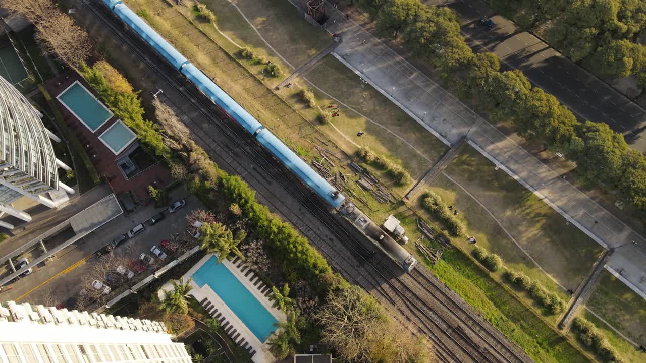 vista aérea de un tren moderno que pasa por una zona urbana, con edificios altos a un lado y camiones en una carretera al otro lado