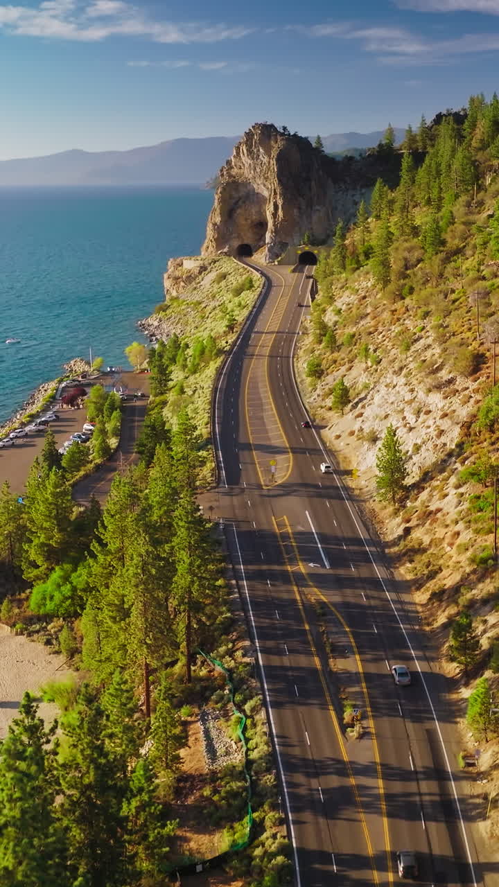 Motorway with tunnel through the mountains. Rocky shore with pine trees near the lake. Blue skies at backdrop. Vertical video