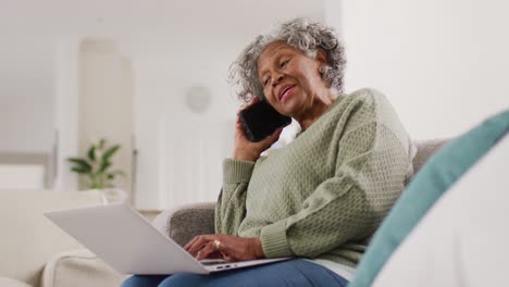 Retrato-De-Una-Anciana-Afroamericana-Sentada-En-Un-Sofá,-Usando-Una-Computadora-Portátil-Y-Hablando-Por-Teléfono-Inteligente
