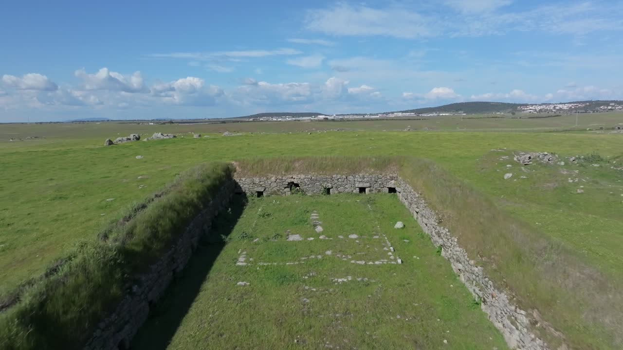 Reverse drone flight over green pastures with surprise reveal of an 18th-century stone structure used for pig farming. The building emerges suddenly from the landscape, surrounded by rural scenery.