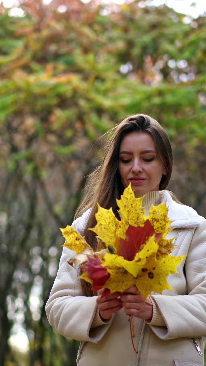 Attractive brunette lady in white jacket walks outdoors with bright leaves in her hands. Woman tosses the leaves into air smiling happily and catching them. Vertical video