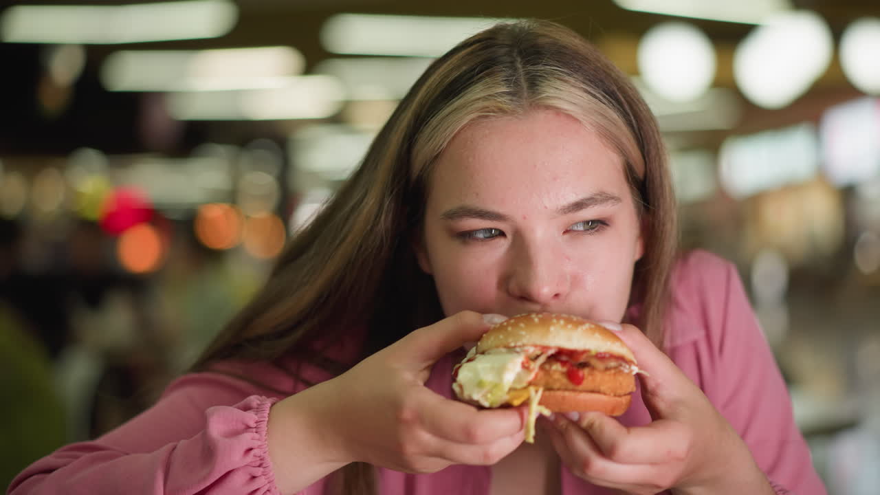 dama de vestido rosa toma un bocado de hamburguesa, masticando lentamente con los ojos bien abiertos, ella parece pensativa, saboreando la comida con enfoque, el fondo está suavemente borroso, con efectos de luz bokeh