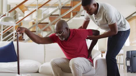 Afroamerikanischer-älterer-Vater-Steht-Von-Der-Couch-Auf-Und-Benutzt-Einen-Stock,-Während-Sein-Jugendlicher-Sohn-Ihm-Hilft