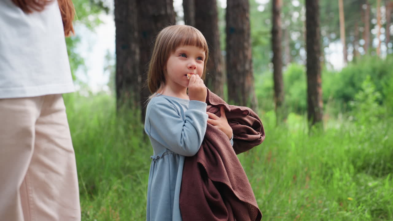 평화로운 숲 속 빈터에서 갈색 담요를 덮고 간식을 먹는 사랑스러운 아기, 녹색 나무 사이로 햇빛이 부드럽게 쏟아지는, 어린 시절의 순수함과 따뜻함을 불러일으키는 부드러운 자연 분위기